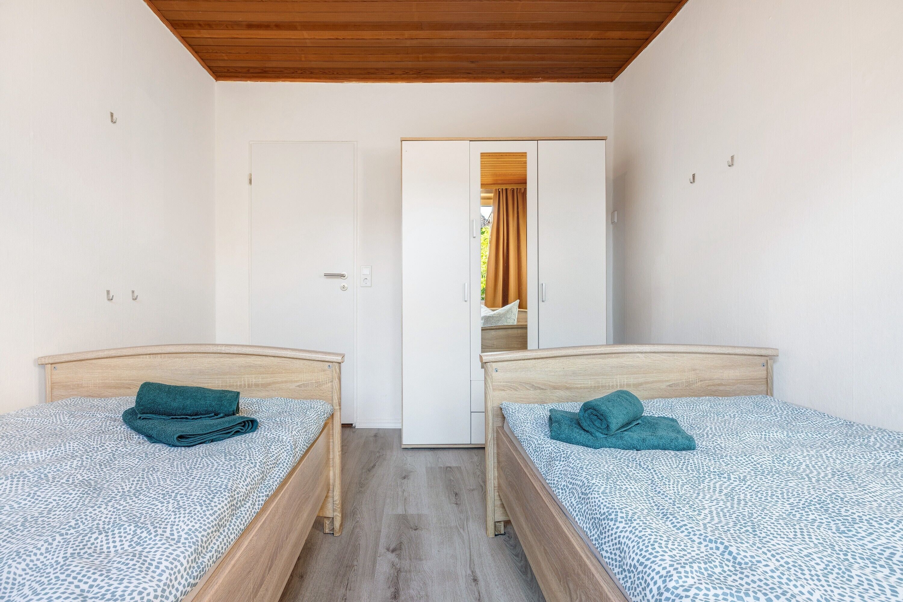A room with two single beds, a wooden ceiling, white walls, and a door leading to another room.