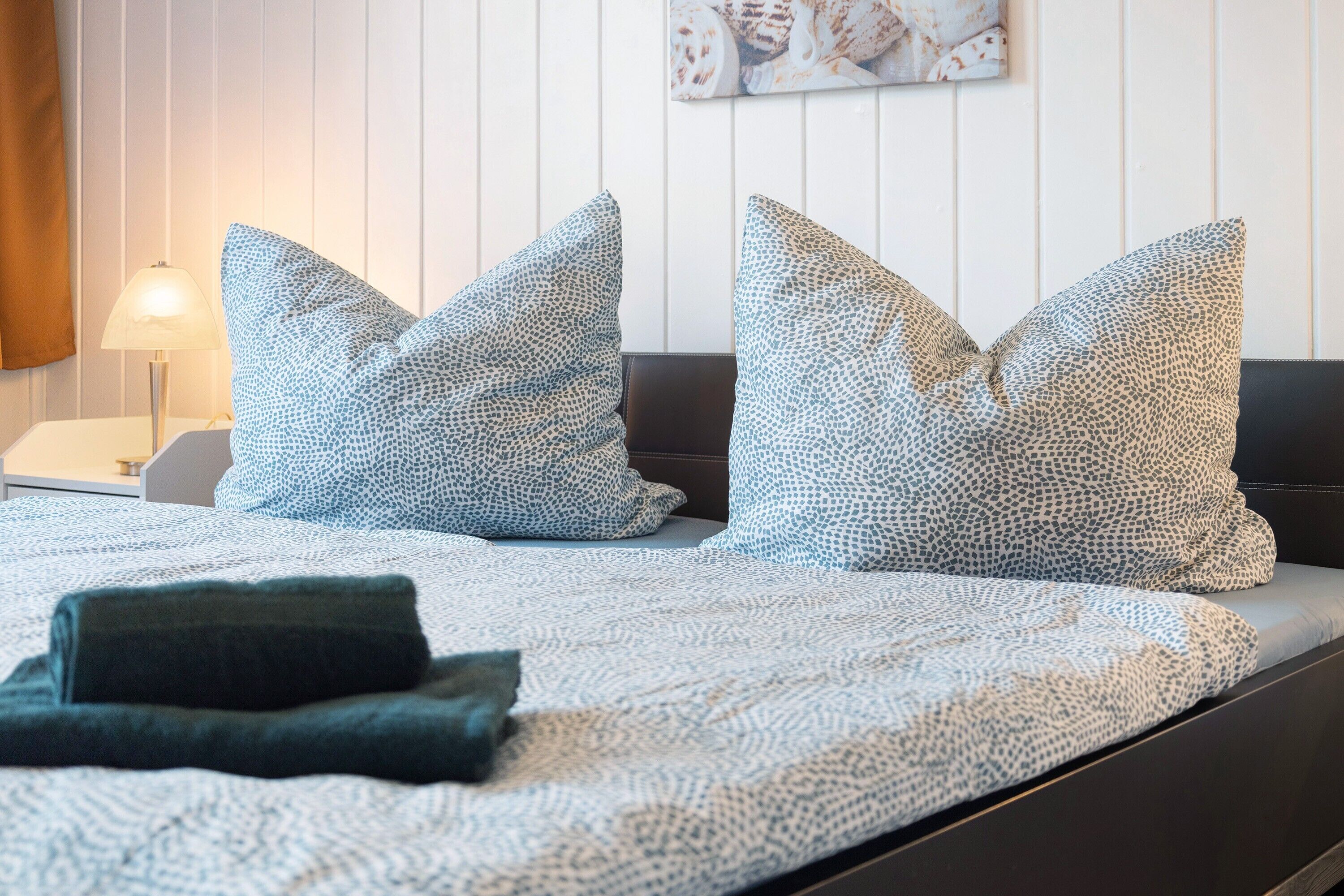 Una cama bien hecha con almohadones estampados, una lámpara de noche y un cuadro en la pared.