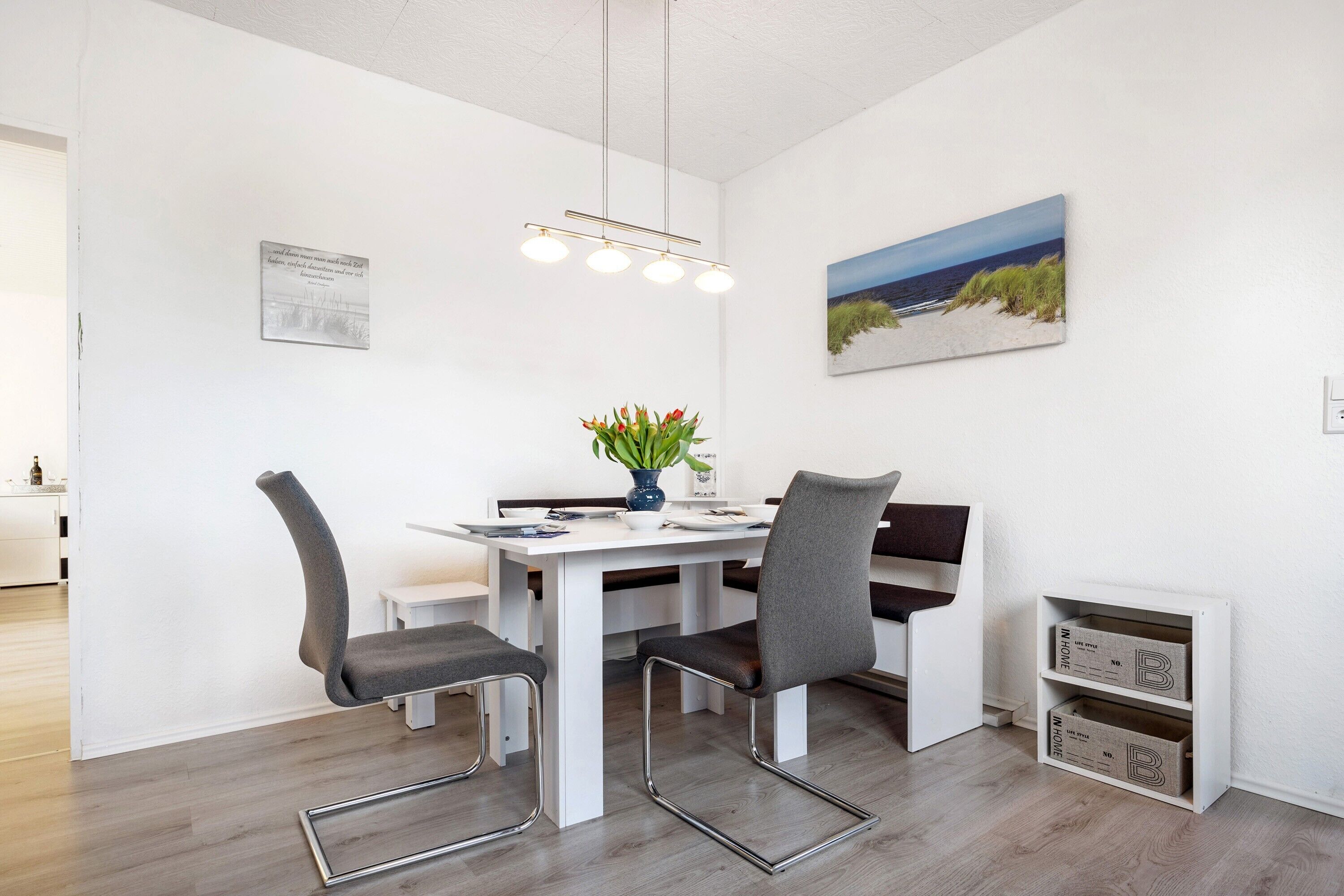 Spiseområde med et hvidt bord, fire stole, en vase med blomster, et billede på væggen og indbyggede skabe.