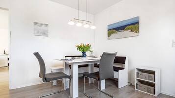 Spiseområde med et hvidt bord, fire stole, en vase med blomster, et billede på væggen og indbyggede skabe.