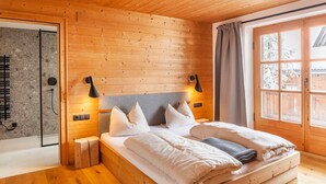 Un dormitorio de cabaña de madera con cama, almohadas, cabecera, mesita de noche, lámpara de pared, ventana con cortinas y zona de ducha.