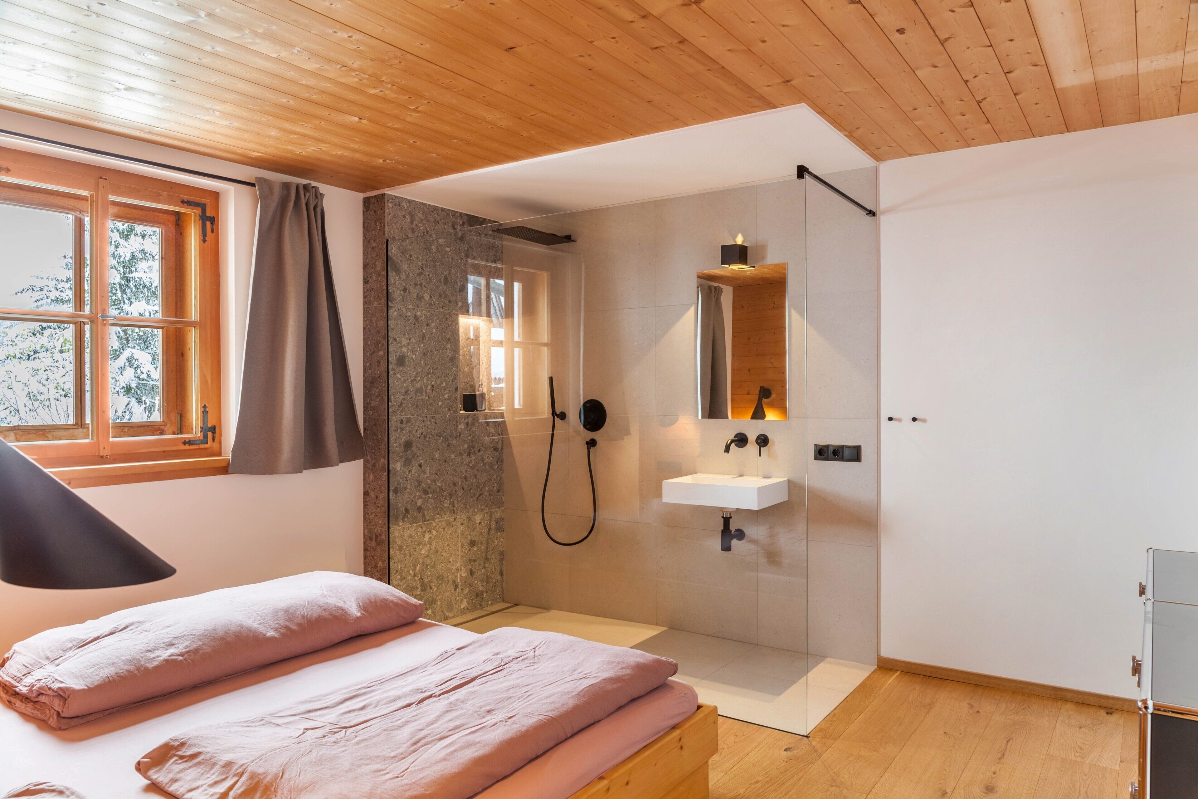 A bedroom with a wooden ceiling, a bed with pink bedding, a window with curtains, a shower area with a glass door, and a white sink.