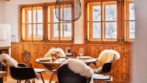 Un interior de madera con una mesa de comedor para cuatro personas, una araña y ventanas grandes que dan a una vista nevada.