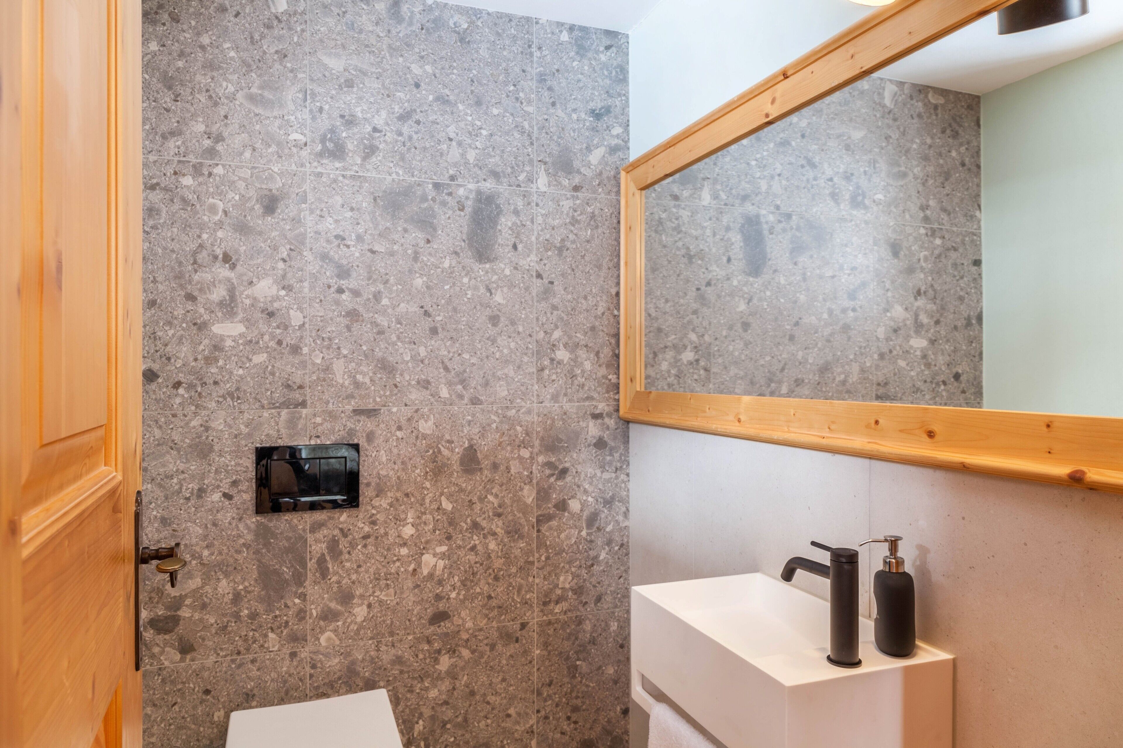 Un baño con pared de piedra, puerta de madera, lavamanos blanco, grifo negro y espejo.