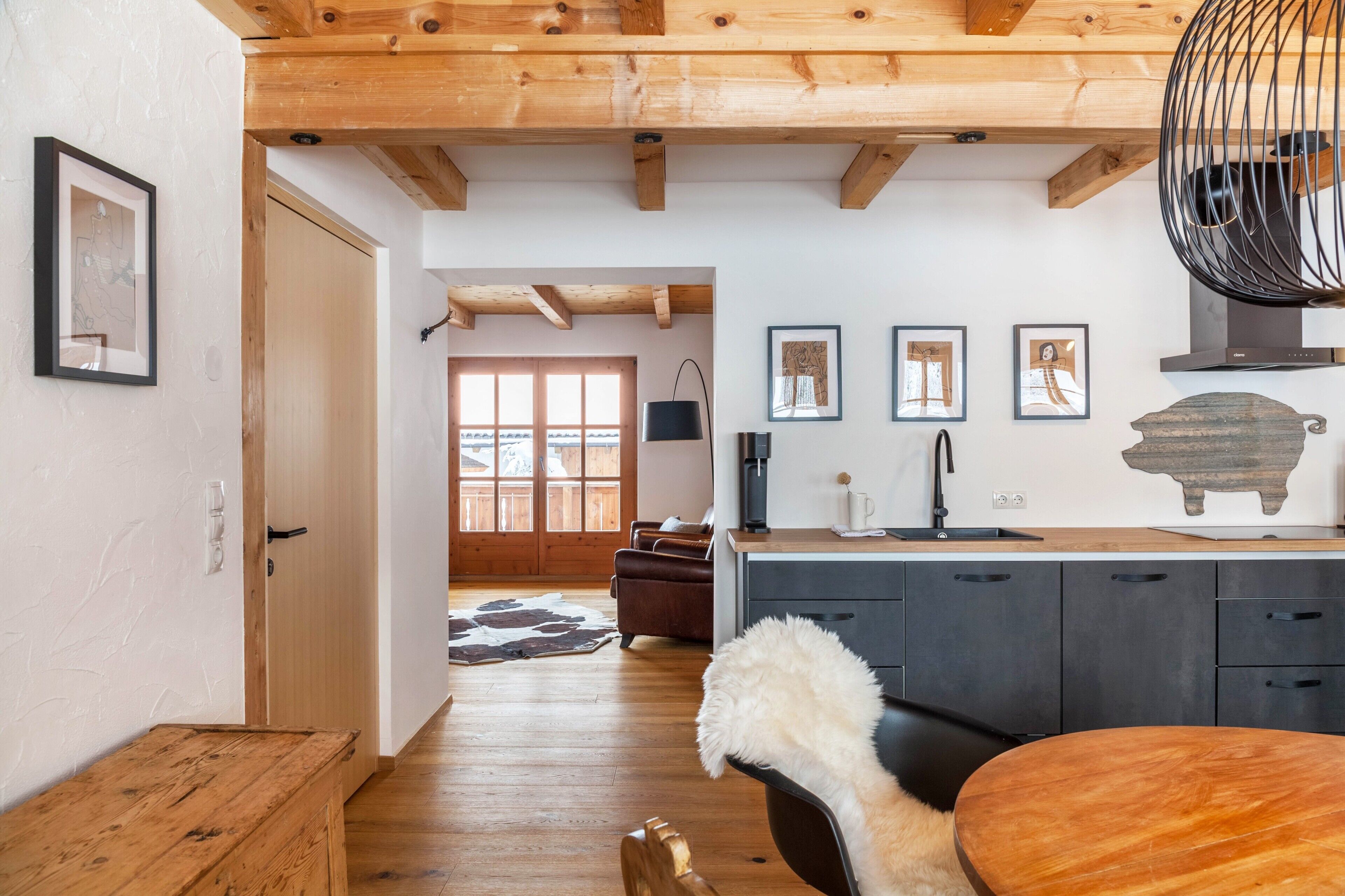 Techo de madera, una mesa de comedor, una silla, un manto de piel y una zona de cocina con armarios y estufa.