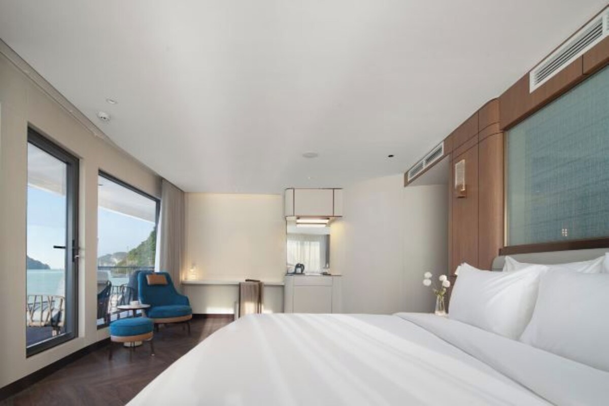 A modern hotel room with a large bed, a desk, and a chair. There is a view of the sea through the windows.