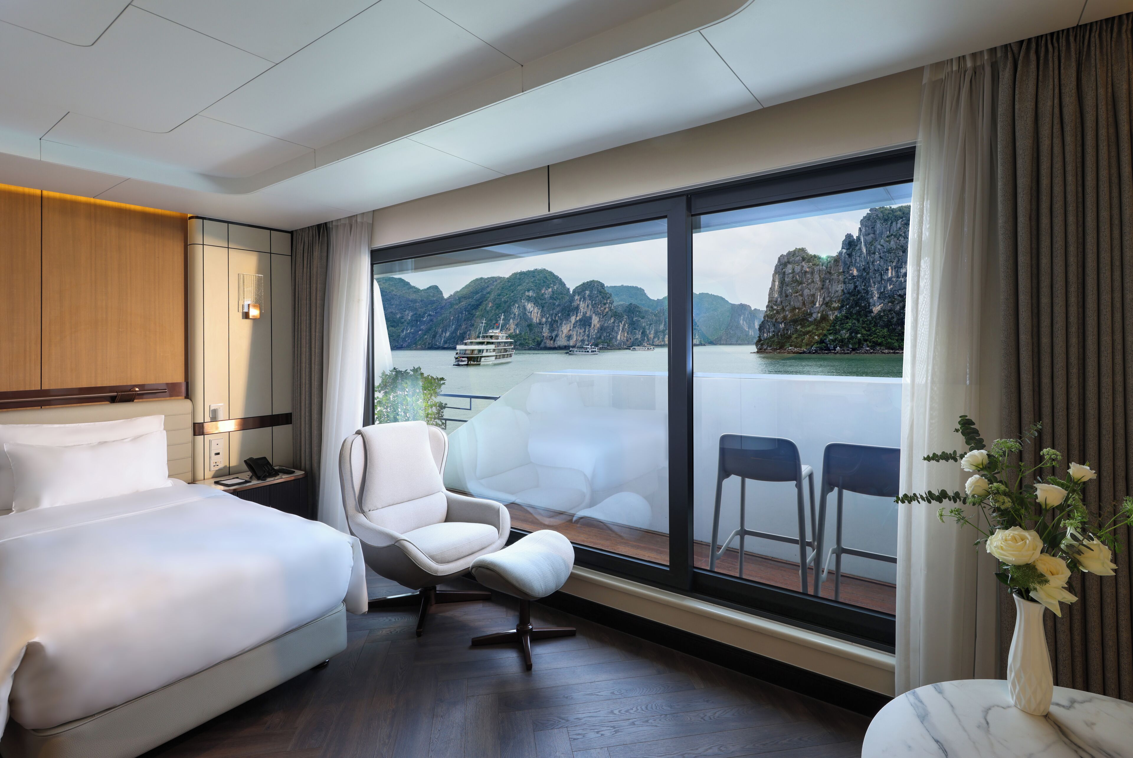 A hotel room with a large window, a bed, a chair, and a vase with flowers.