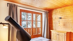 Una habitación de cabaña de madera con una cama, una ventana con cortinas, una lámpara fijada a la pared y un armario de madera.