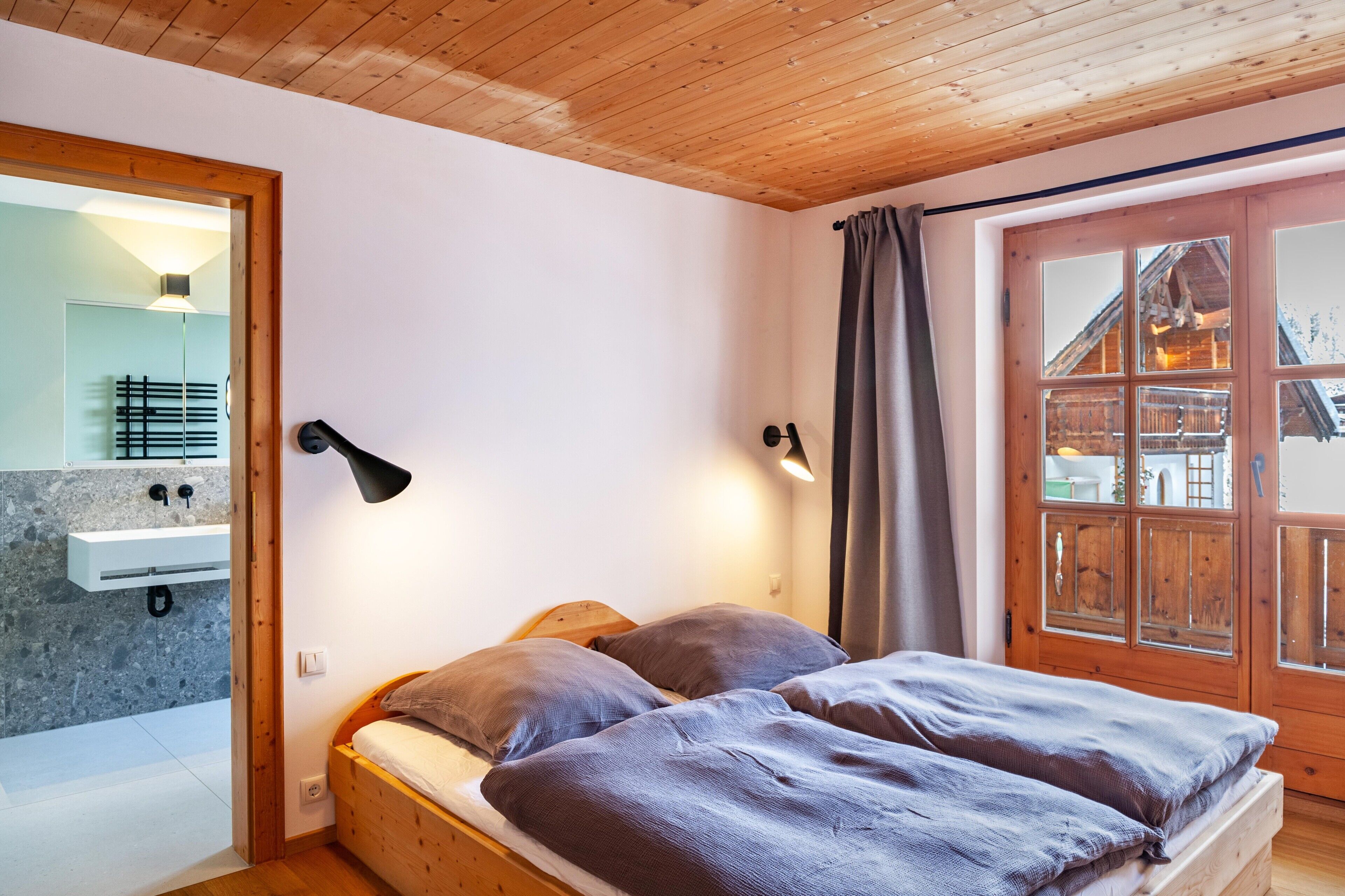 A wooden ceiling, a bed with two pillows, a wooden door with glass panels, and a bathroom with a stone sink.