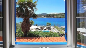 Une vue par la fenêtre sur une zone côtière, avec une mer bleue et limpide, un palmier et un toit recouvert de tuiles.