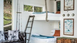 Un lit superposé avec une échelle, un canapé agrémenté de coussins à motifs de palmiers et une malle en bois.