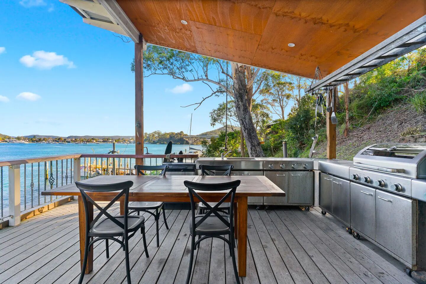 An outdoor deck with a wooden table and chairs, a grill, and a view of the water.
