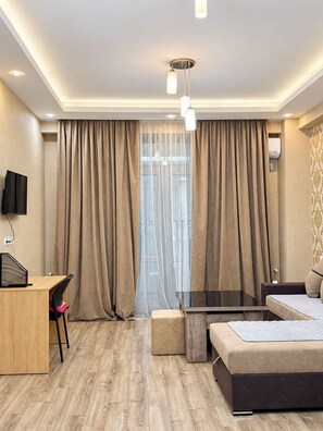 A modern hotel room with a wooden desk, a glass table, a sofa, and a large window with curtains.