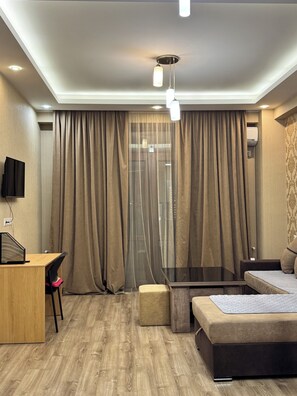 A modern hotel room with a wooden desk, a sofa, a glass-top coffee table, and a large window with curtains. - Saburtalo, Jhonberi 2 (T'bilisi)