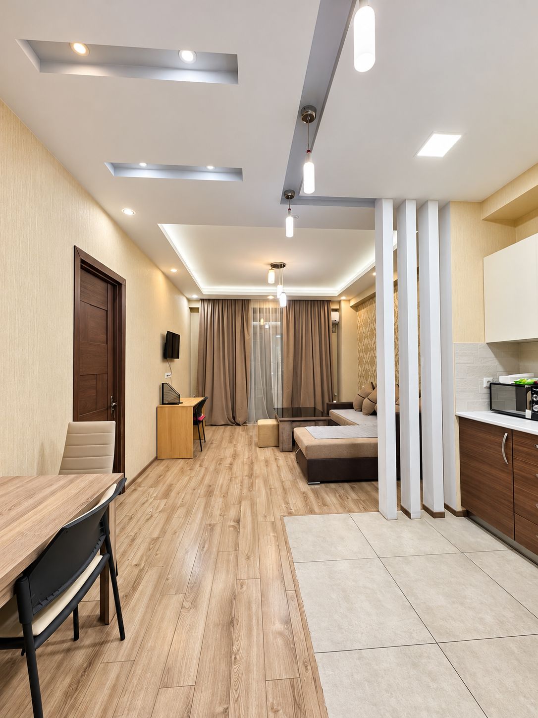 A modern hotel room with a wooden floor, a dining area, a kitchenette, and a large window with curtains.