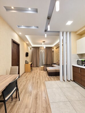 A modern hotel room with a wooden floor, a dining area, a kitchenette, and a large window with curtains.