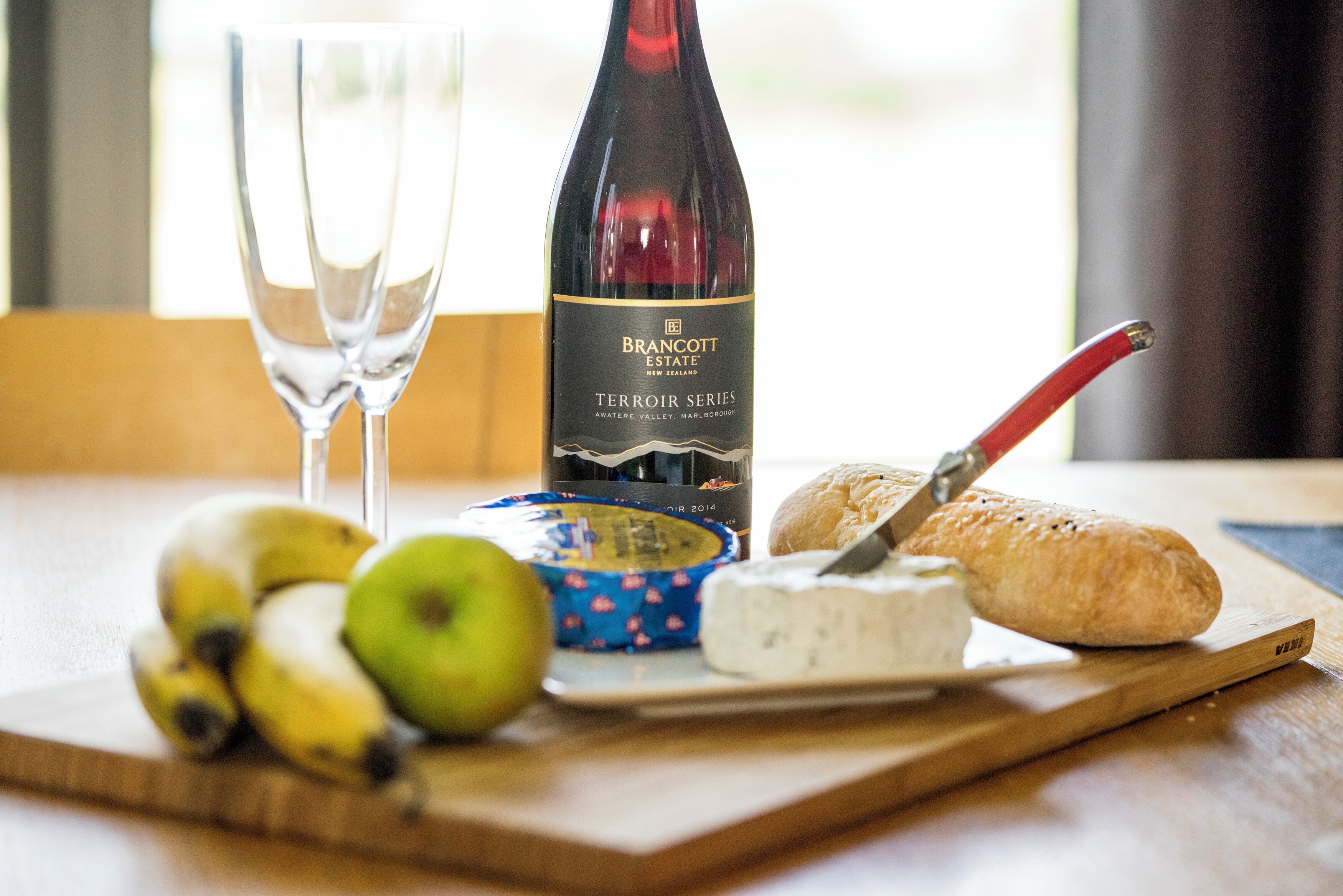 A table with a bottle of wine, two glasses, a loaf of bread, a cheese, and bananas.