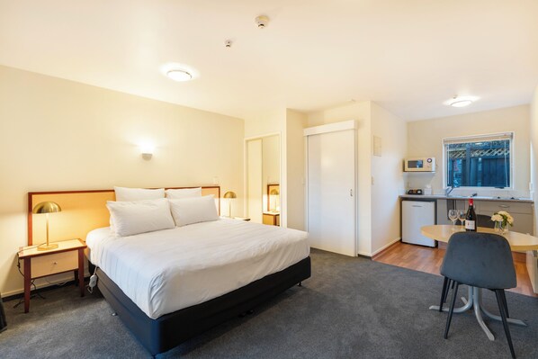 Habitación de hotel con cama, mesitas de noche, escritorio con silla, kitchenette pequeña y ventana.