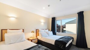 A hotel room with two beds, a nightstand with a lamp, and a large window with curtains. - Daffodil Motel (Christchurch)