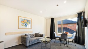 A modern living room with a grey sofa, a glass coffee table, and a dining area with a small round table and blue chairs. - Daffodil Motel (Christchurch)
