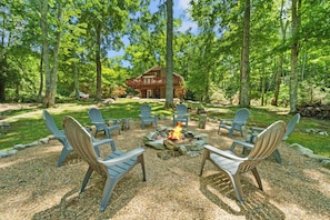 Un chalet doté d’un foyer entouré de fauteuils Adirondack, situé dans un espace boisé avec une grande maison en arrière-plan.