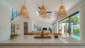 A modern kitchen with a large island, hanging wicker pendant lights, and a view of a swimming pool. - Puri Senang - Modern 4BR Pool Villa · Central Berawa · 6 min to Beach & Finns (Canggu)