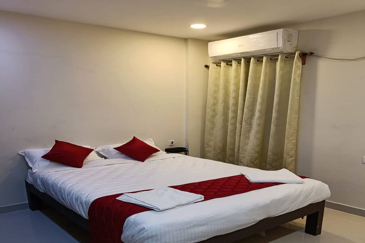 A double bed with white and red bedding, a red throw blanket, and a white towel. There is a nightstand with a lamp, a window with beige curtains, and an air conditioning unit above the window.