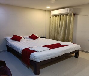 A double bed with white and red bedding, a red throw blanket, and a white towel. There is a nightstand with a lamp, a window with beige curtains, and an air conditioning unit above the window. - Sai Residency (Puducherry)