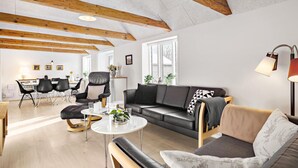 A modern living room with a wooden ceiling, a black leather sofa, a round coffee table, and a dining area with chairs and a table. - 3 bedroom stunning home in Løkken (Løkken)