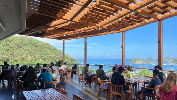 An outdoor dining area with wooden tables and chairs, overlooking a scenic view of the sea and hills. - ZASGO HOTEL (Labuan Bajo)