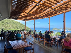 An outdoor dining area with wooden tables and chairs, overlooking a scenic view of the sea and hills. - ZASGO HOTEL (Labuan Bajo)
