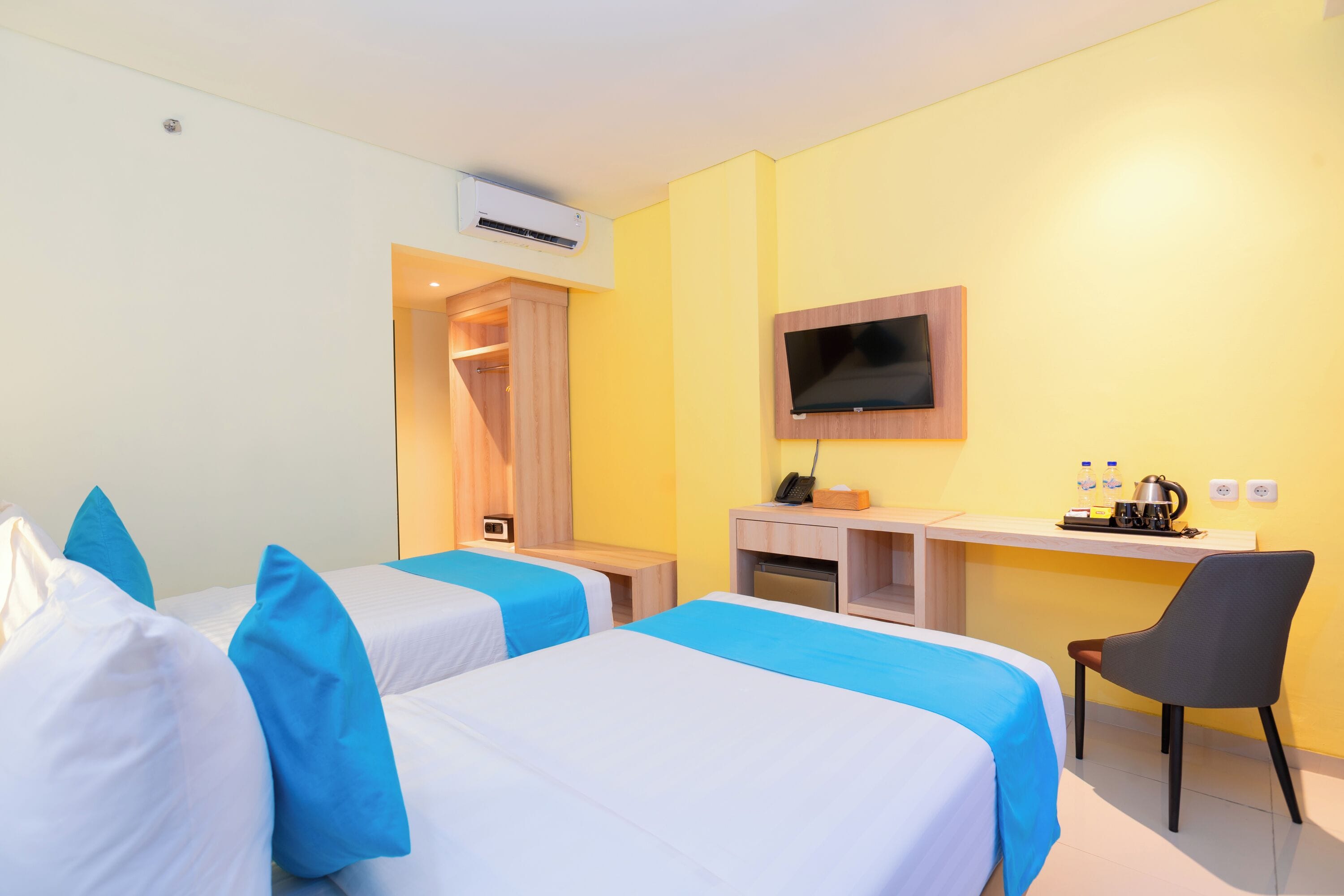 A hotel room with two beds, a desk, a chair, a TV, and an air conditioning unit.