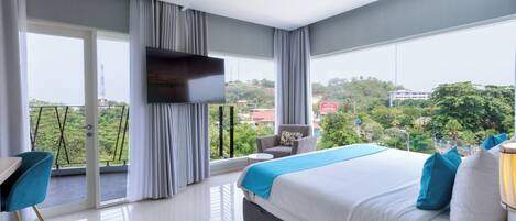 A modern hotel room with a large bed, a TV, a balcony with curtains, and a view of the city.