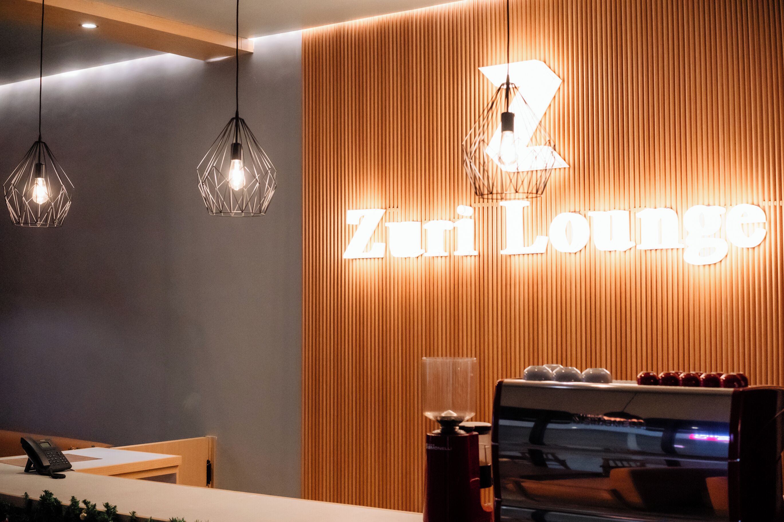 Un espace d’accueil avec un mur en bois, une enseigne « Zurli Lounge » et des luminaires suspendus.