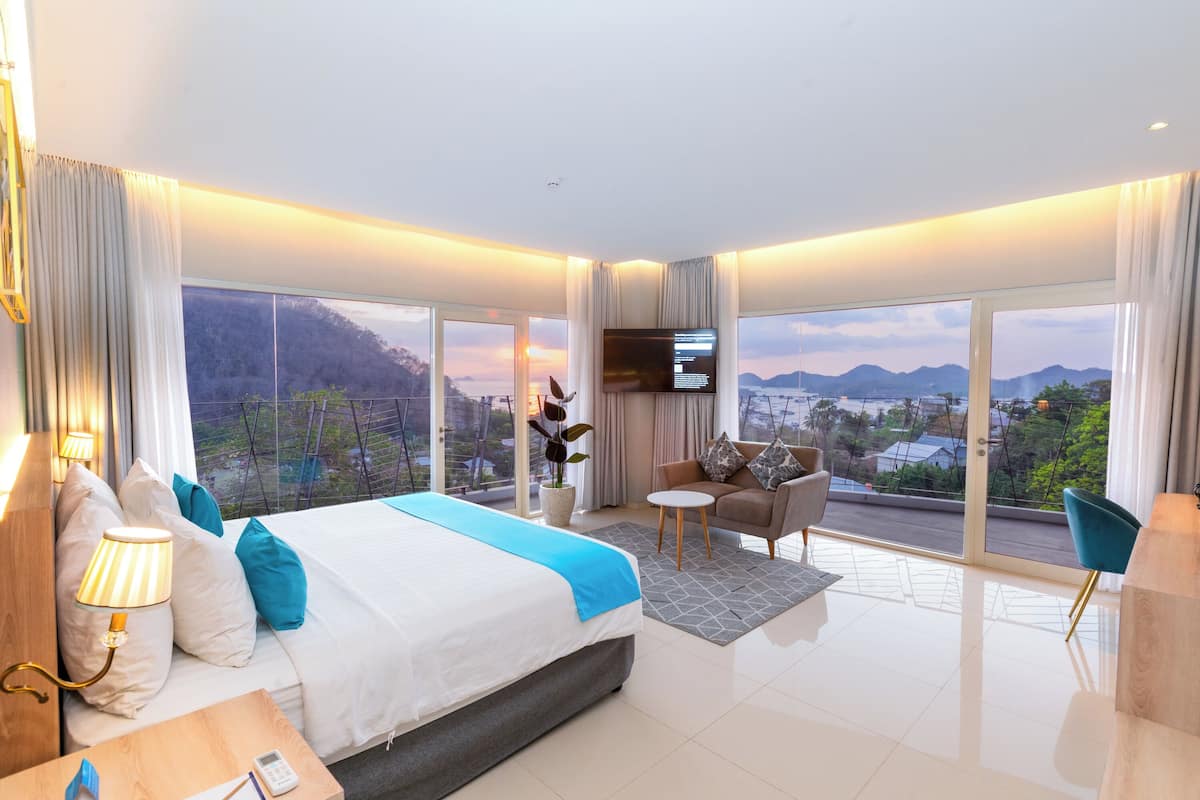 A modern hotel room with a large bed, a TV, a sofa, and a view of the city.