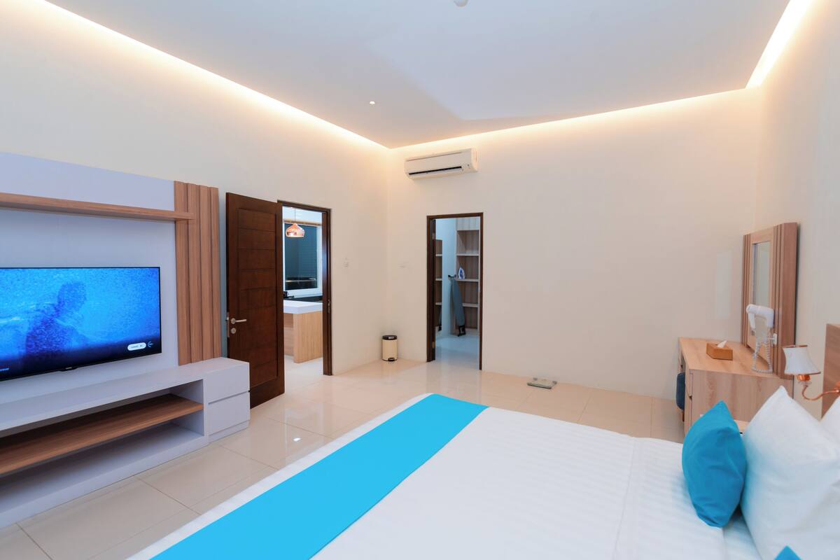 A modern hotel room with a large flat-screen TV, a bed with blue and white bedding, a wooden nightstand, and a desk with a lamp.