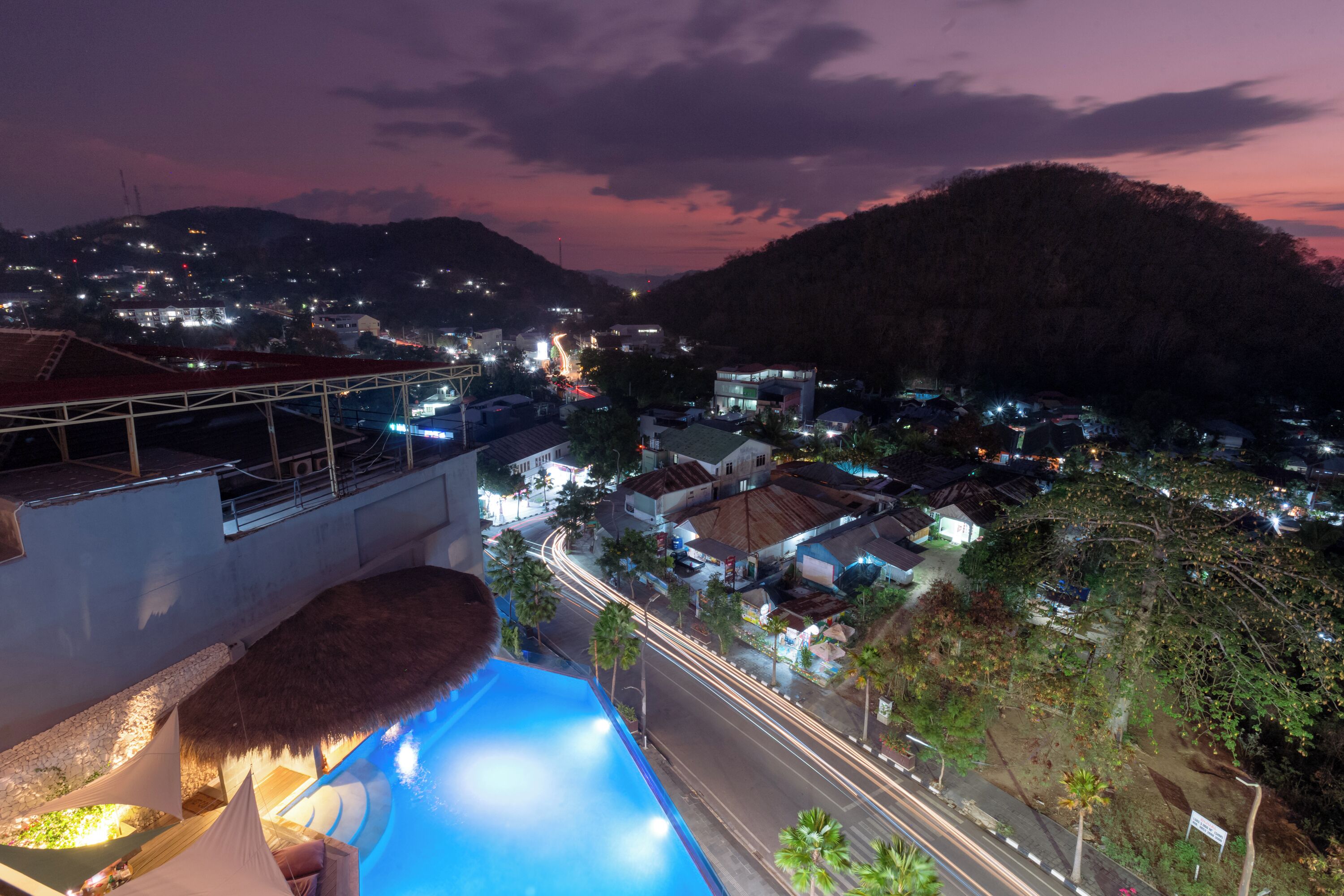 Vista nocturna de un complejo turístico con piscina, construcción de techo de paja y vistas del pueblo y las colinas circundantes.