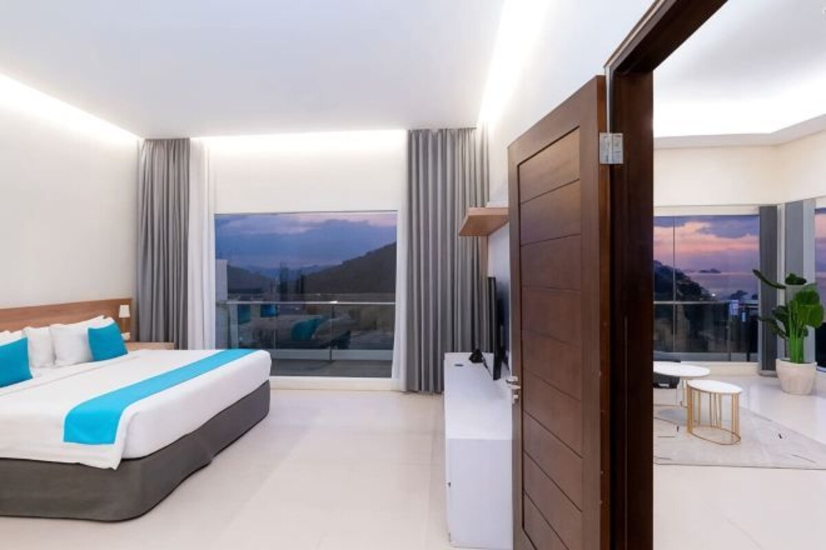 A modern hotel room with a large bed, a balcony with a view, and a small seating area.