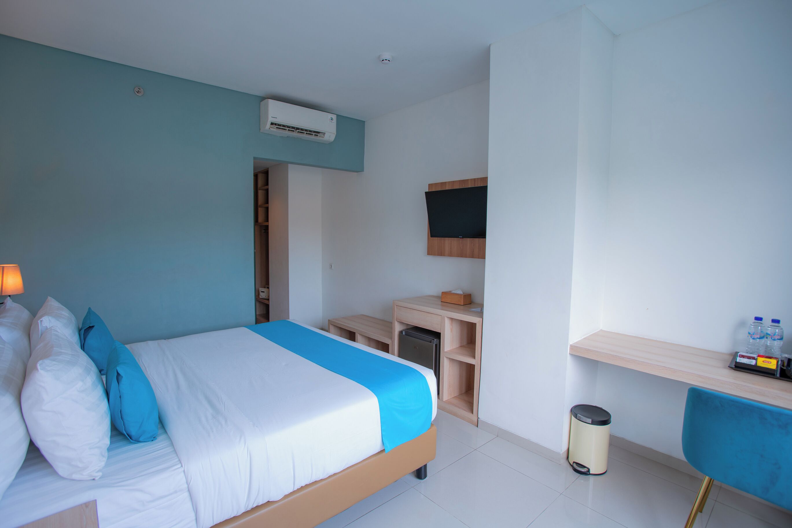 A hotel room with a bed, a desk, a chair, a TV, and a trash bin.