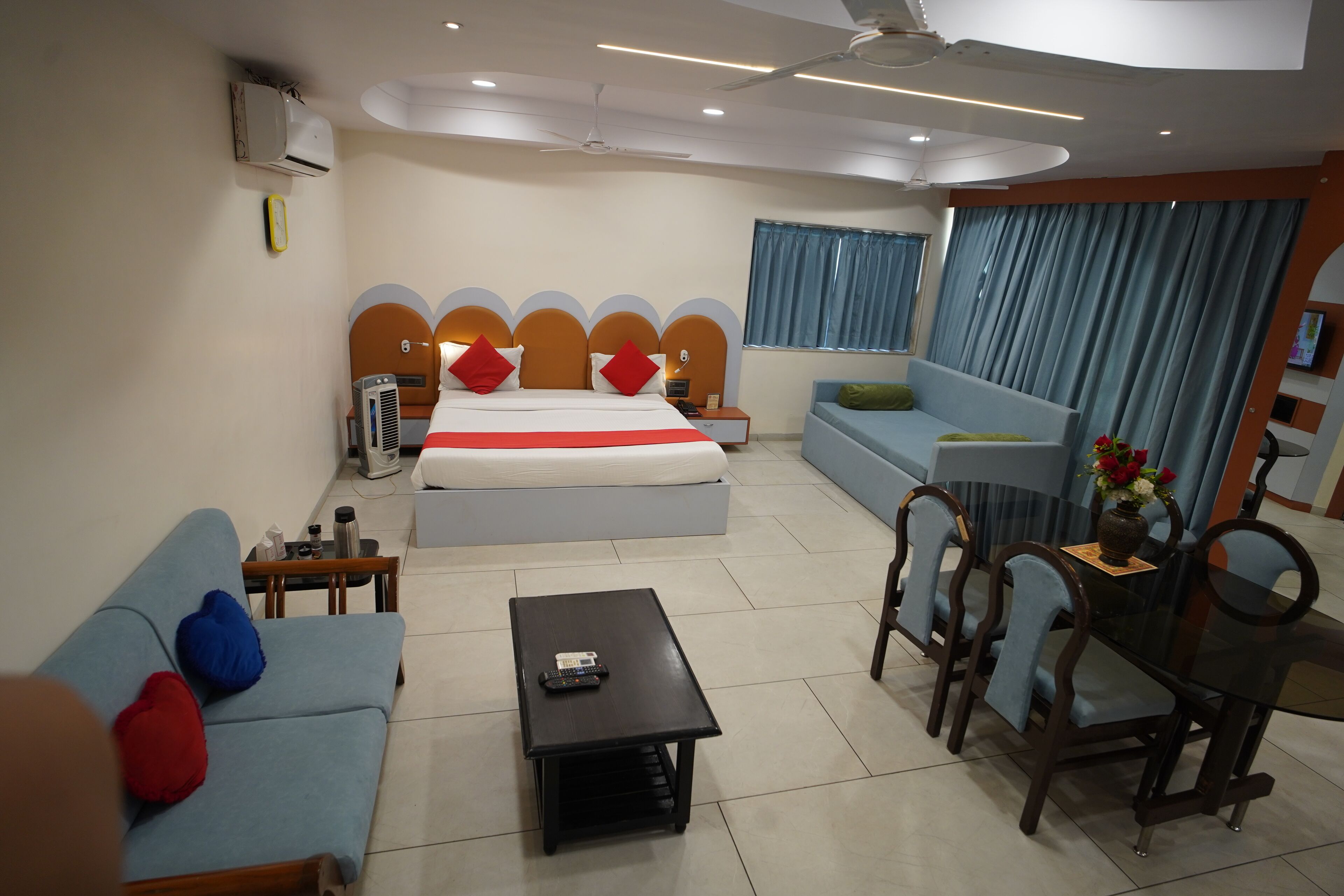 A modern hotel room with a large bed, a sofa, a dining table, and a ceiling fan.
