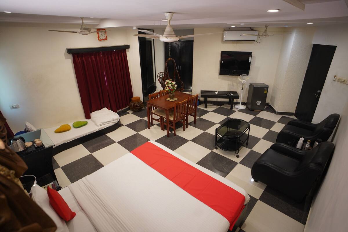 A hotel room with a checkered floor, a bed with a red and white bedspread, a dining table with chairs, a sofa, a TV, and a ceiling fan.