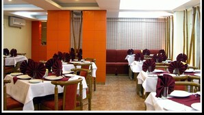 A restaurant with tables set for dining, a red cushioned seating area, and a large window with curtains. - Hotel Gokul (Gandhidham)