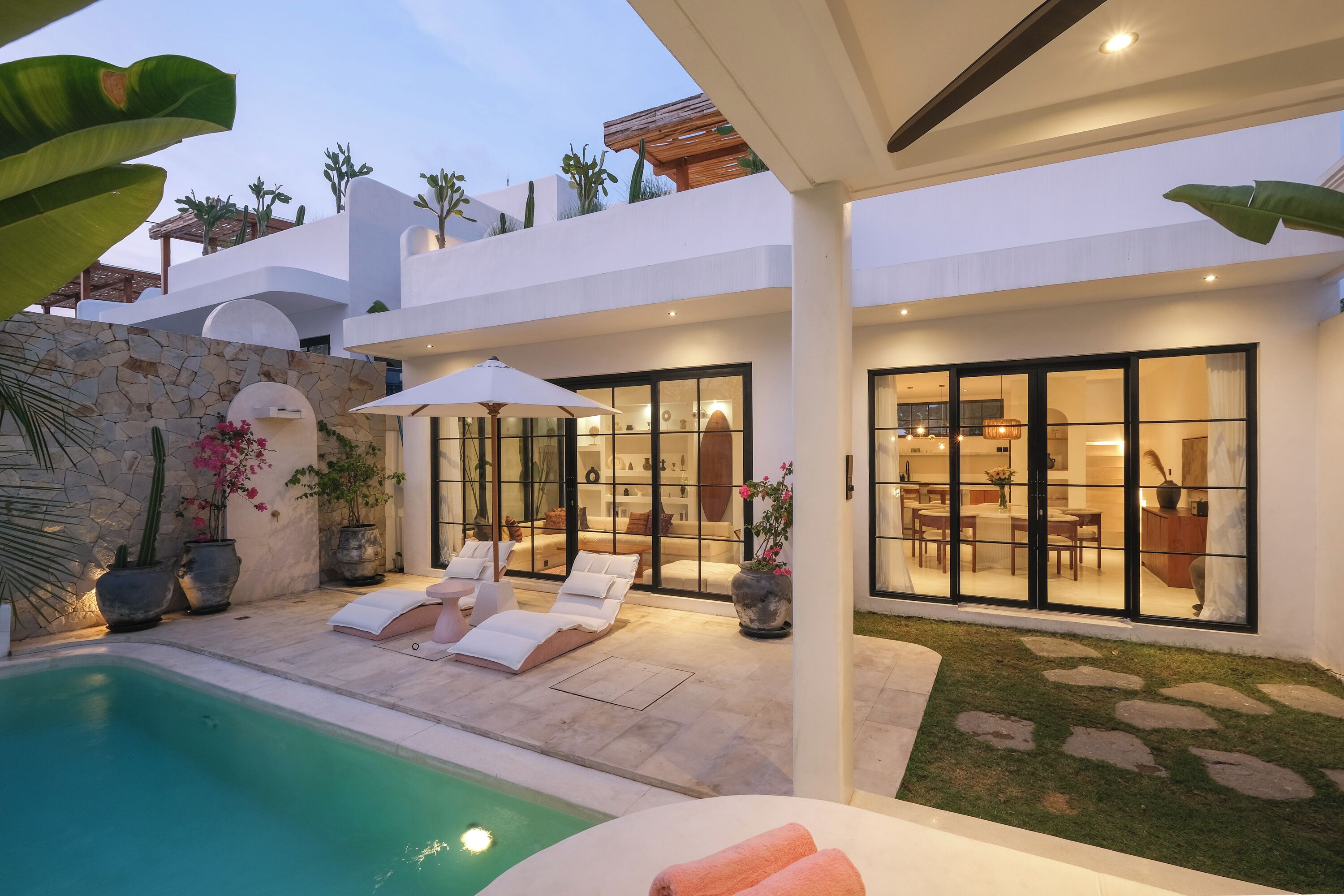 A modern house with a swimming pool, lounge chairs, and a well-lit interior.