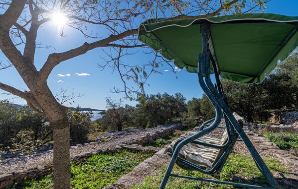 Uma cadeira de metal com toldo verde, colocada sob uma árvore, com vista para uma paisagem rochosa e um corpo d'água ao longe.