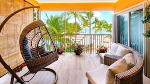 A balcony with a wicker chair, wicker sofa, and a view of palm trees and the ocean.