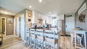 A modern kitchen with white cabinetry, a central island with bar stools, and a refrigerator. - Balcony + Pools + Pickleball + Tennis | Beachfront Port A | Blue Water Bliss (Port Aransas)