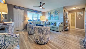 A living room with a wooden floor, a ceiling fan, a couch, two ottomans, and a door labeled "Port Aransas". - Balcony + Pools + Pickleball + Tennis | Beachfront Port A | Blue Water Bliss (Port Aransas)