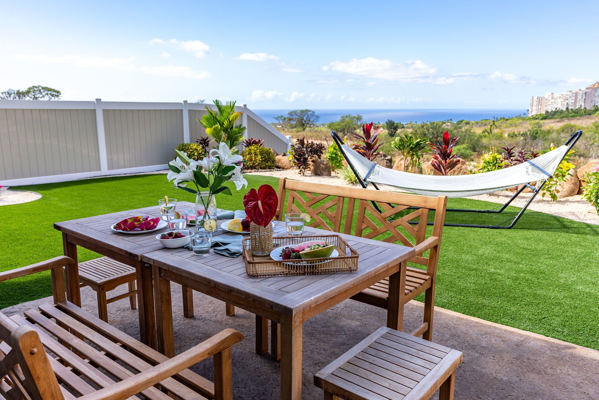 Una mesa de madera al aire libre con vistas al océano, rodeada de sillas y una hamaca.