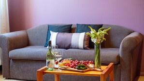 Un salon avec un canapé gris, une table basse en bois, une bouteille de vin blanc, des verres, une assiette de raisins, des concombres et un vase rempli de fleurs.