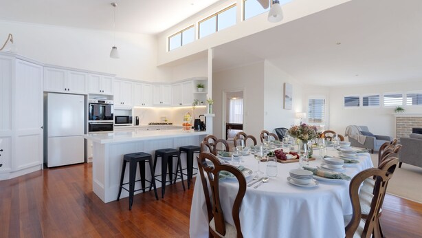 Une cuisine moderne avec des meubles blancs, un îlot central doté de tabourets et un coin repas aménagé pour six personnes, avec une nappe blanche.